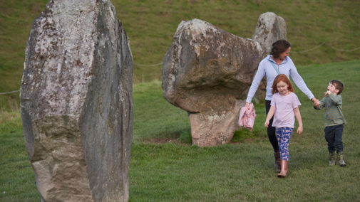 A woman and two children are walking hand-in-hand between large standing stones at the Avebury stone circle, with green grass and the henge bank in the background.